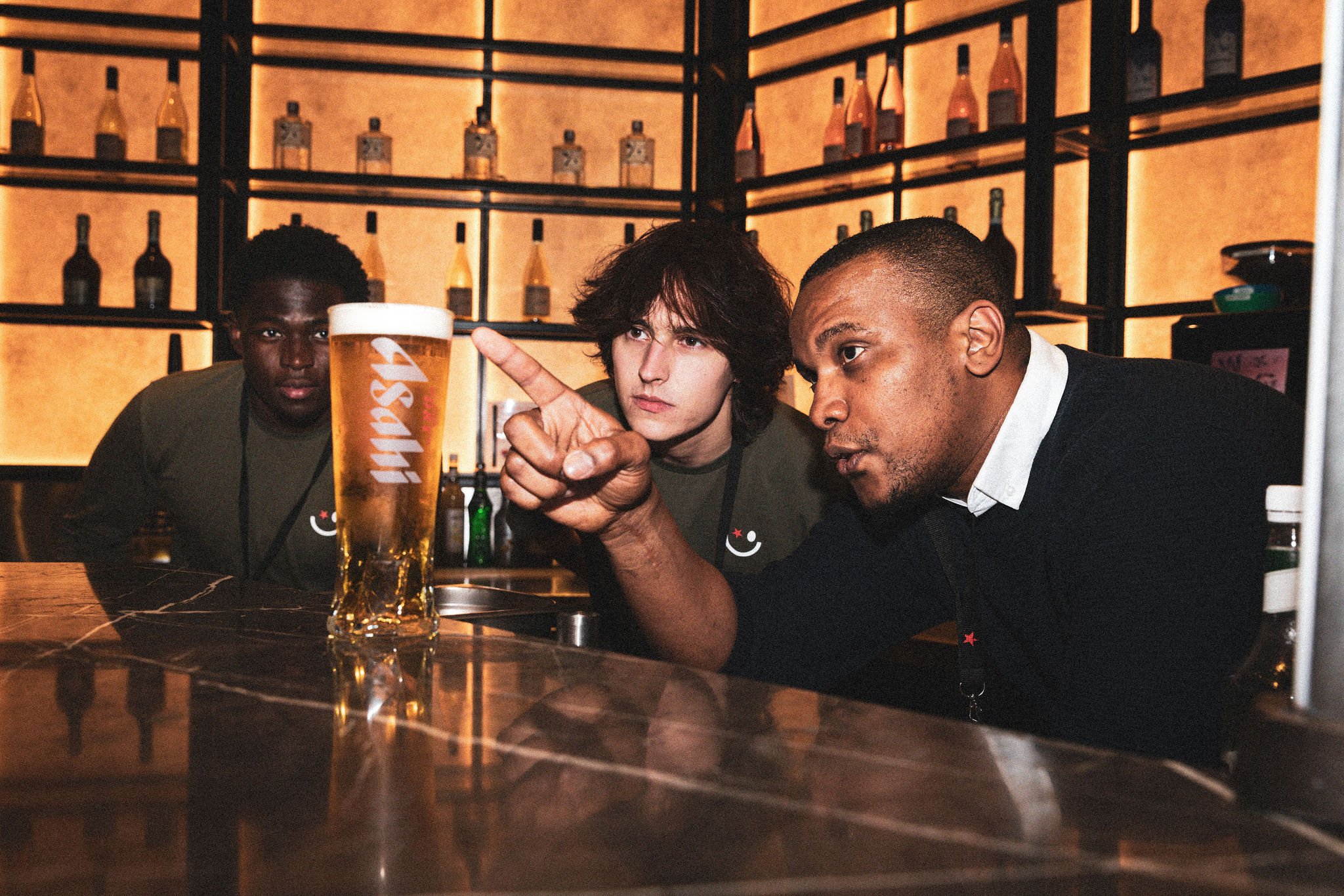 Three people at a bar looking at a beer.