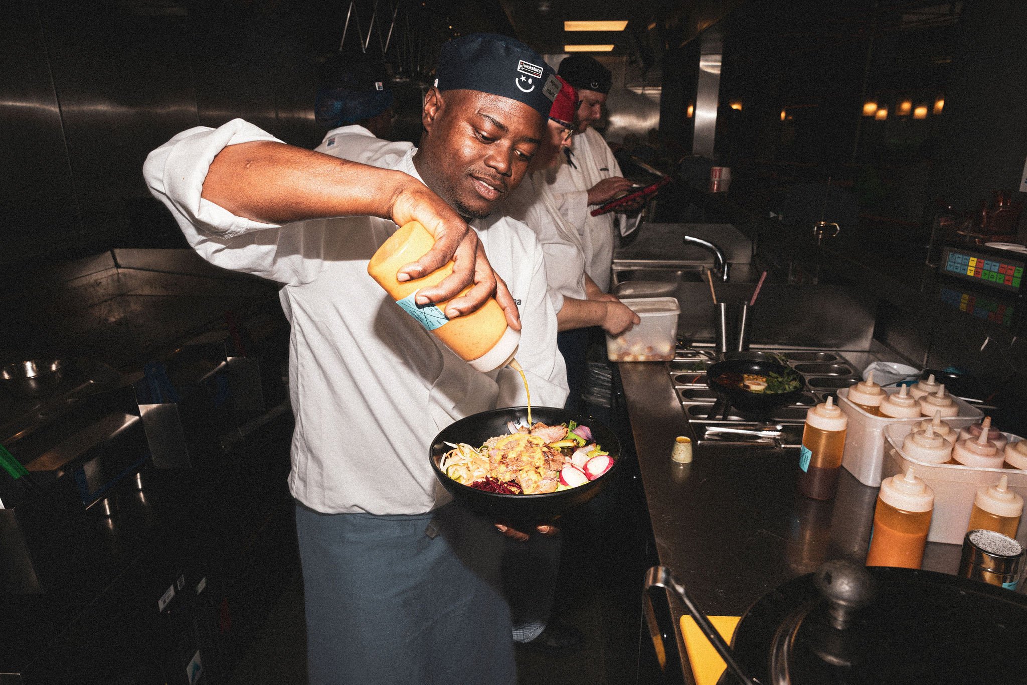Chef in a kitchen pouring sauce on a dish. Other chefs in the background.