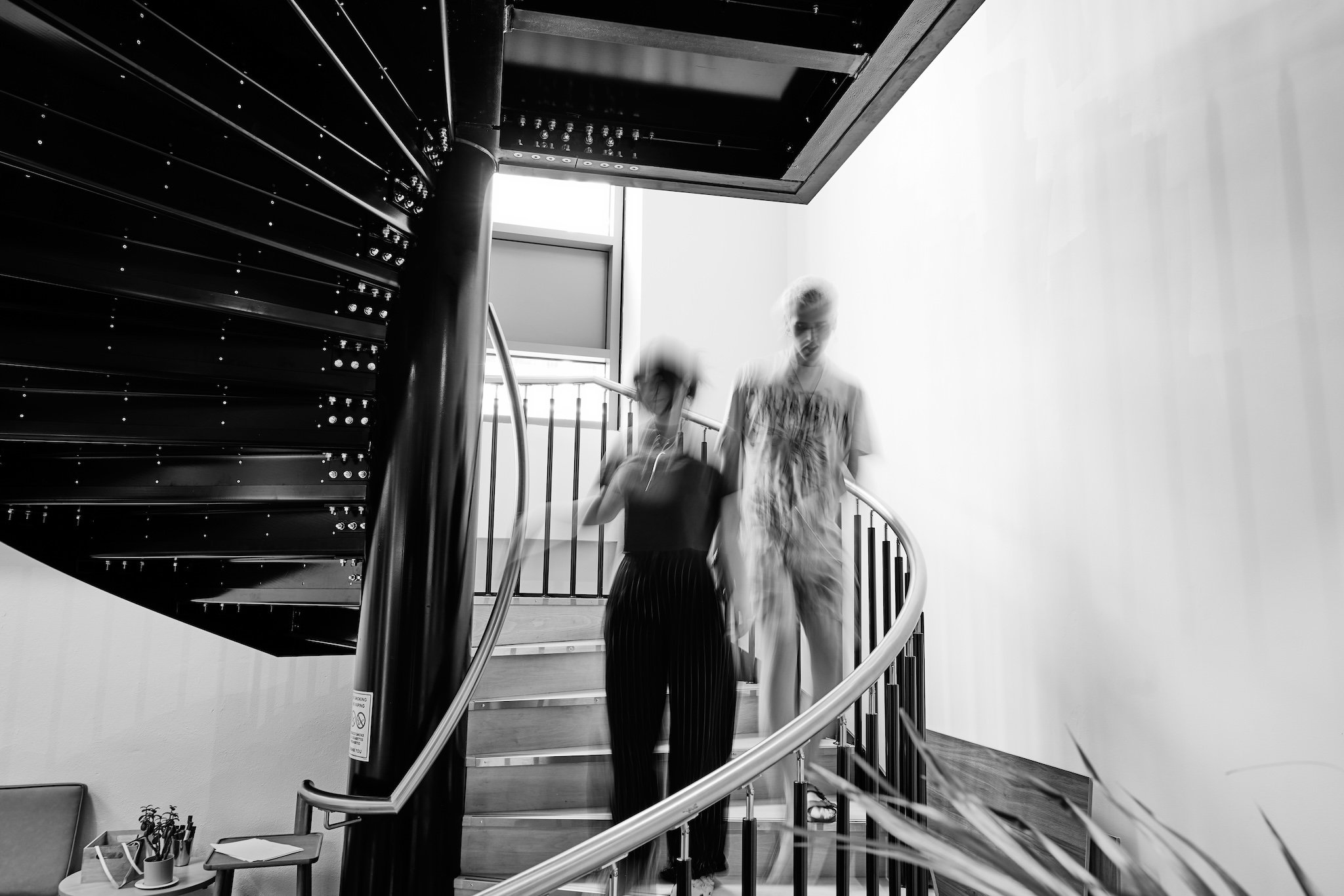 Black and white photo of spiral staircase with two blurry figures descending.