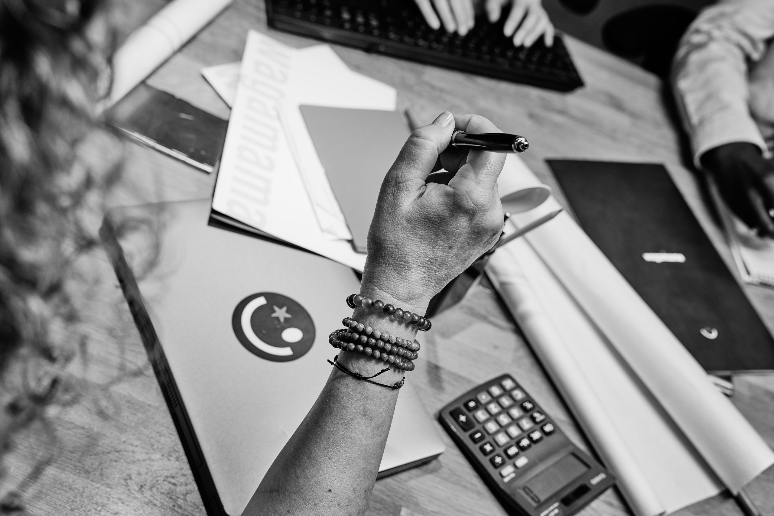Person's hand holding a pen at a desk with papers, calculator, and other items.