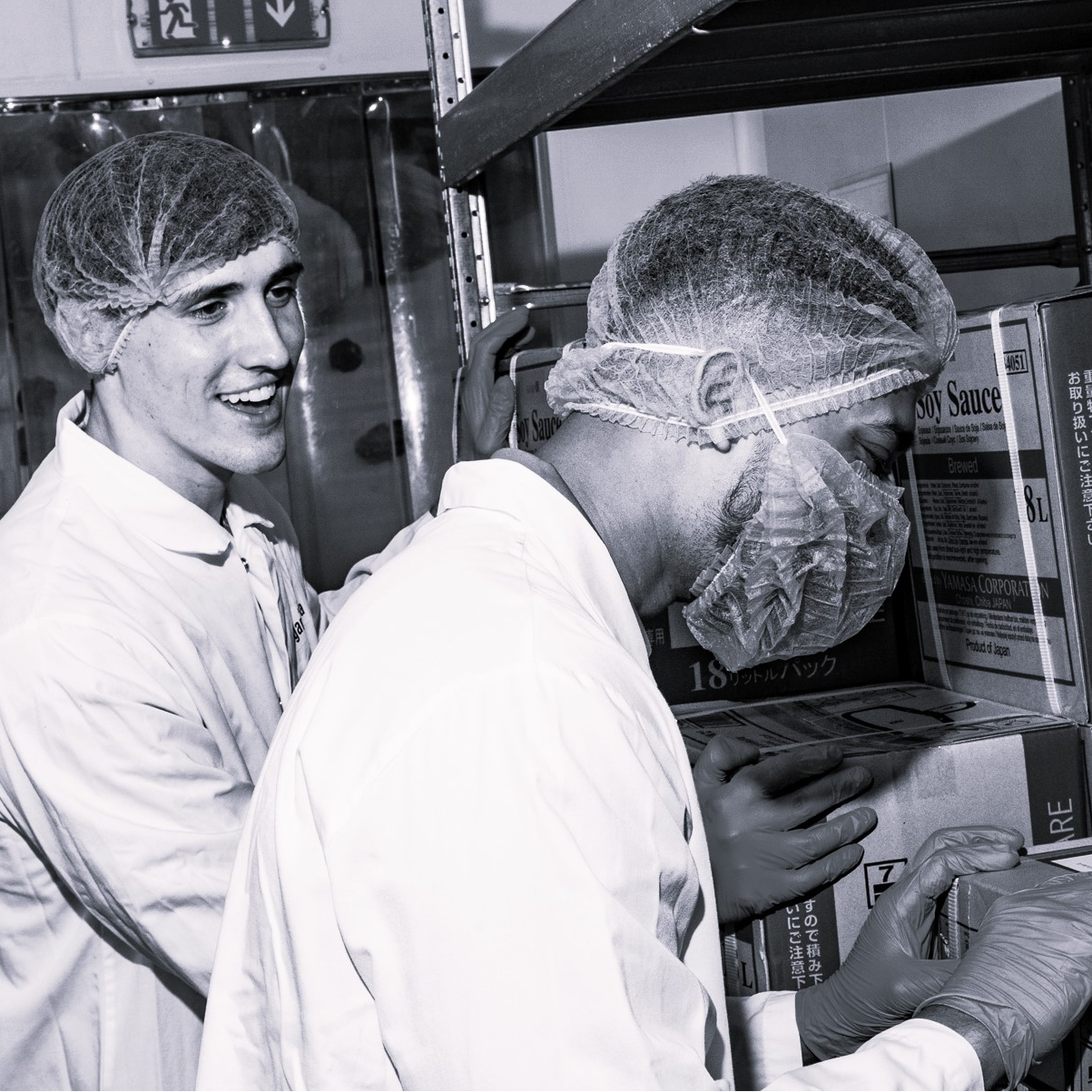Two men in lab coats and hair nets near boxes.