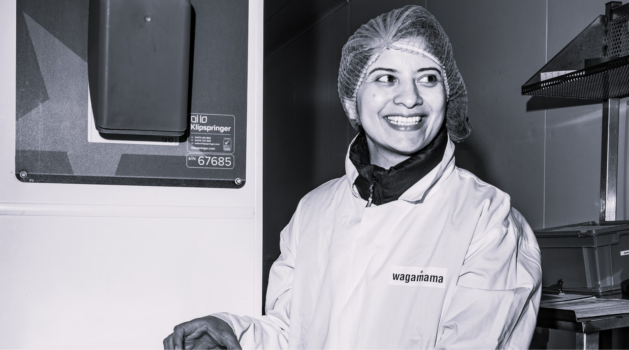 Woman in hairnet and coat with "wagamama" logo, smiling in a kitchen.