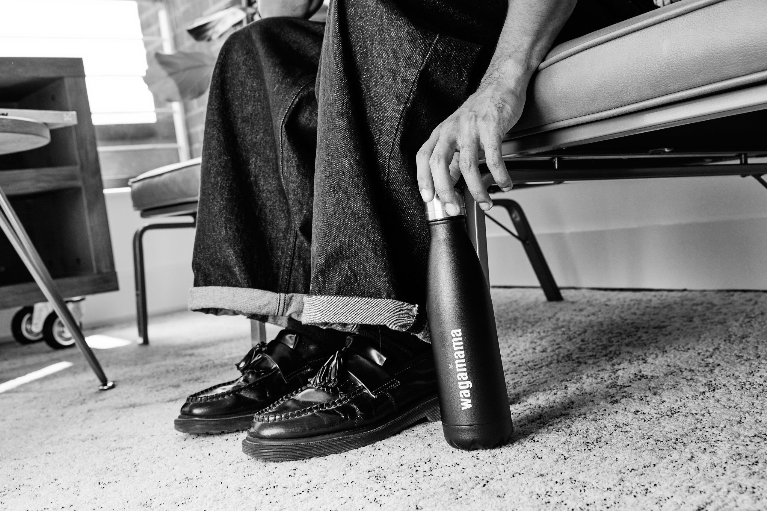 Person holding a black Wagamama bottle, wearing loafers.