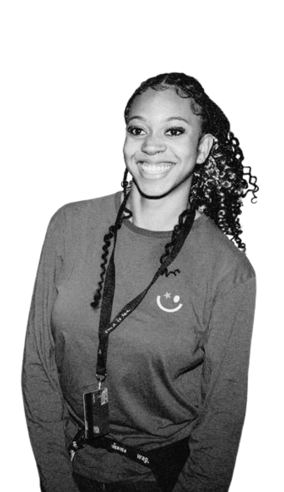 A smiling young woman with curly hair wearing a long-sleeved shirt and a lanyard against white.