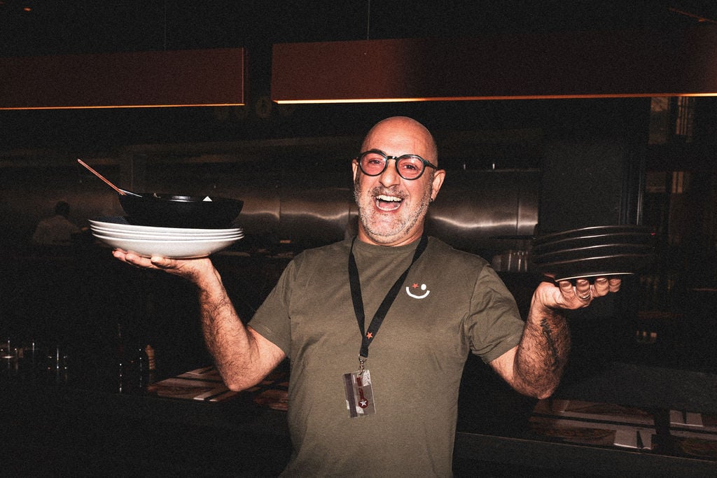 A smiling bald man with glasses in a green shirt holding stacks of empty plates in a restaurant.