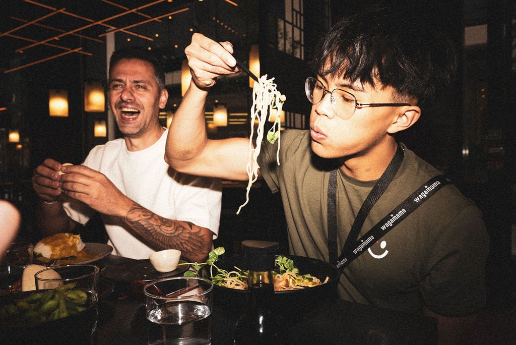 A young man eats noodles with chopsticks while a laughing man sits beside him at a restaurant.
