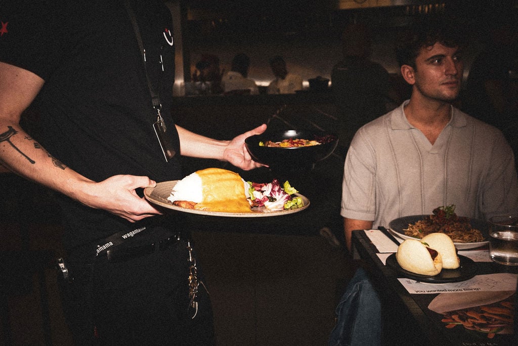 A waiter in a black uniform serving a plate of rice and curry to a customer at a restaurant.