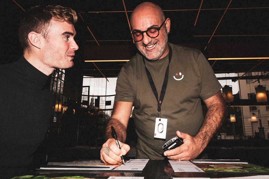 A smiling man with glasses signs documents for a younger man in a dimly lit, modern interior.