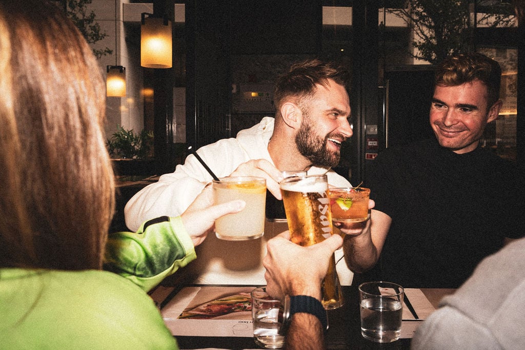 A group of friends laughing while toasting with glasses of beer and cocktails in a dimly lit bar.