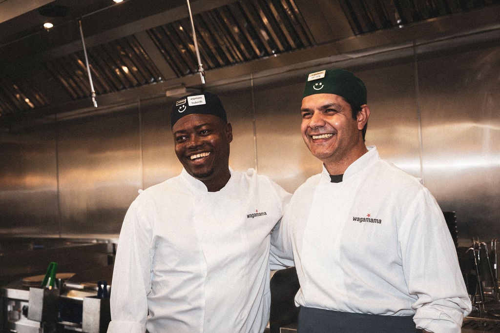 Two smiling Wagamama chefs in white uniforms and branded caps standing in a commercial kitchen.