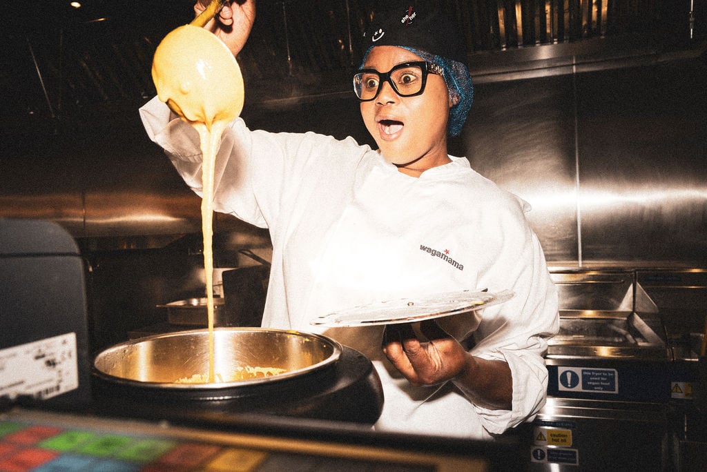 A surprised Wagamama chef in uniform lifts a ladle of yellow sauce over a metal pot in a kitchen.