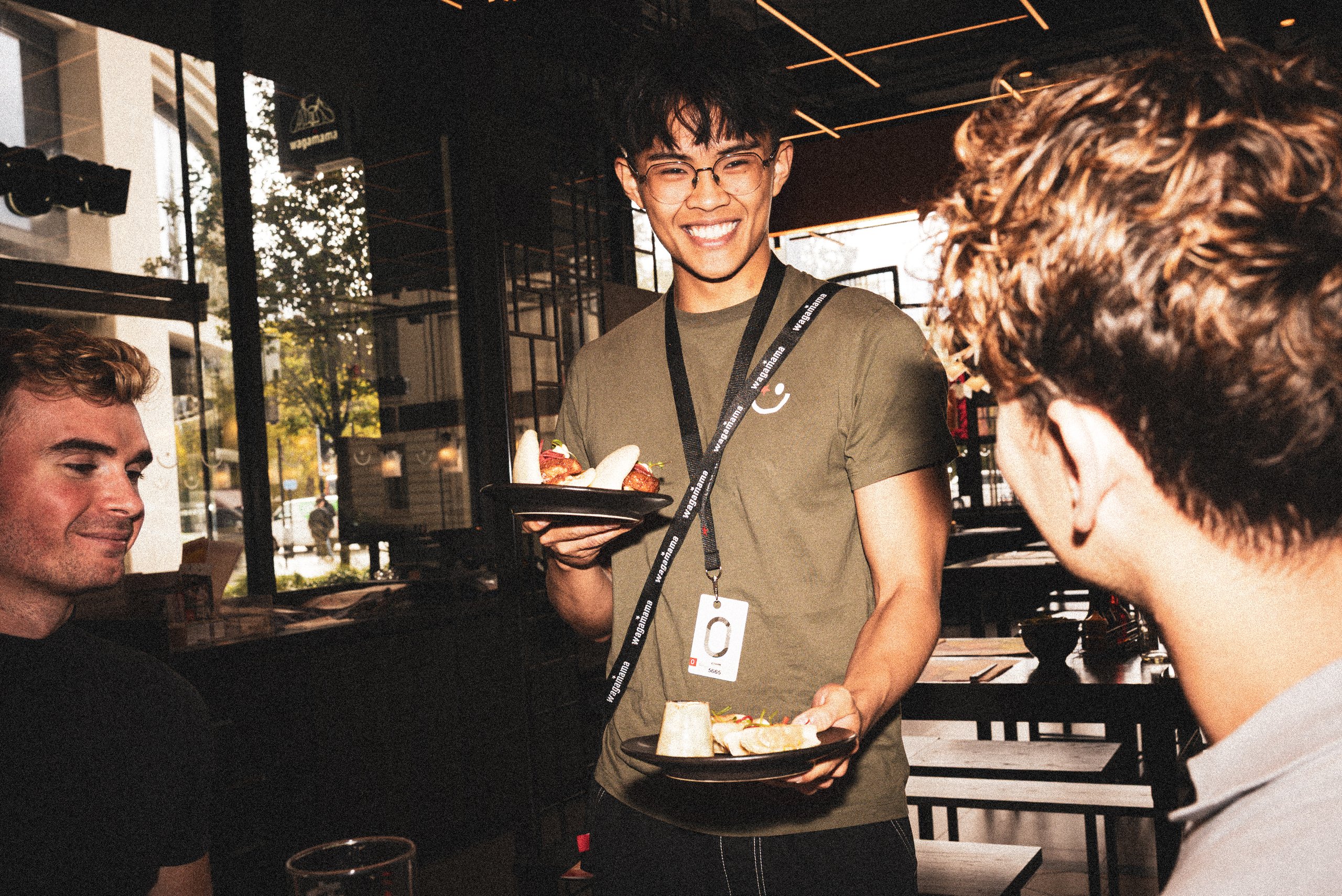 A smiling Wagamama server in a green t-shirt serving plates of food to two customers in a restaurant.