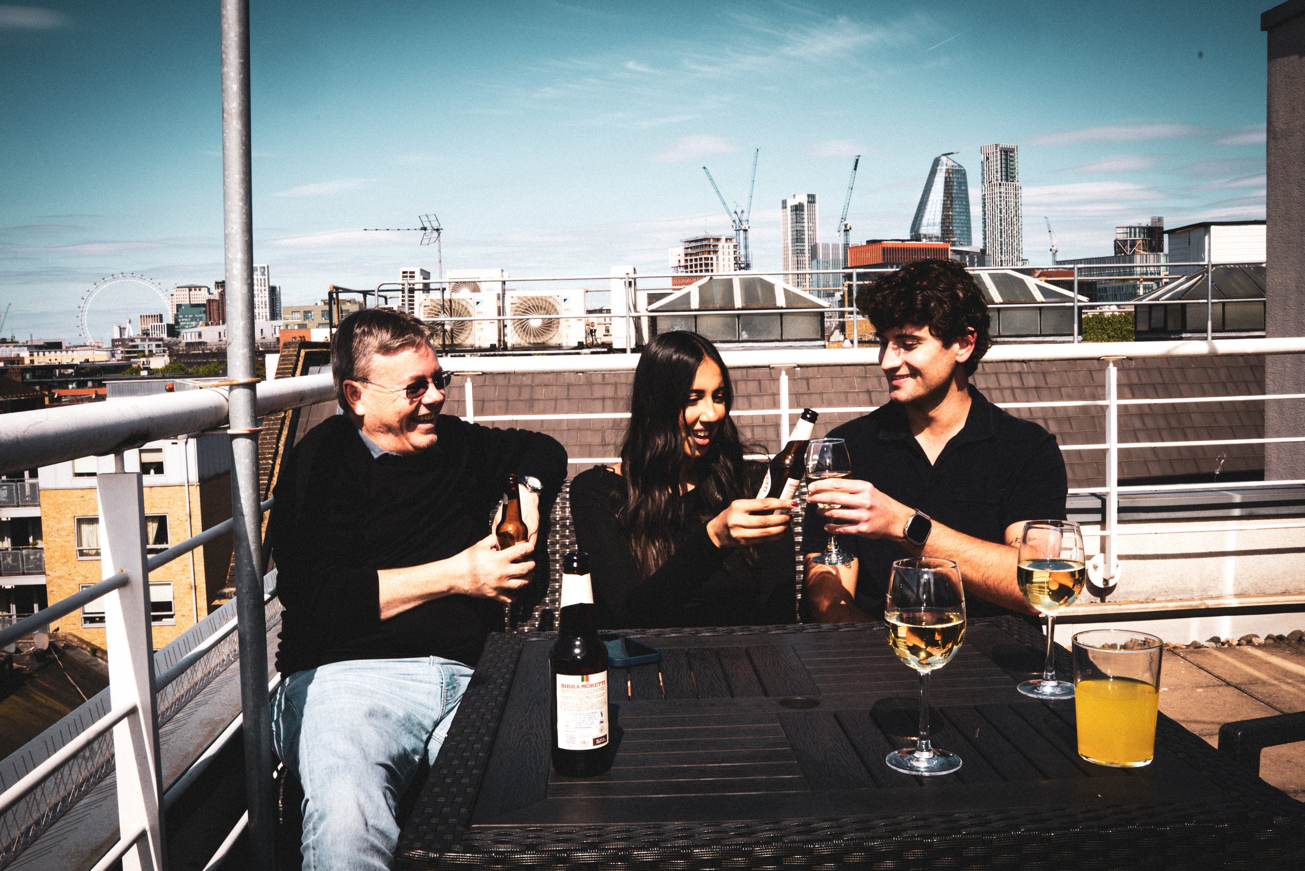 Three people sitting at an outdoor table on a sunny rooftop terrace with the London skyline behind.