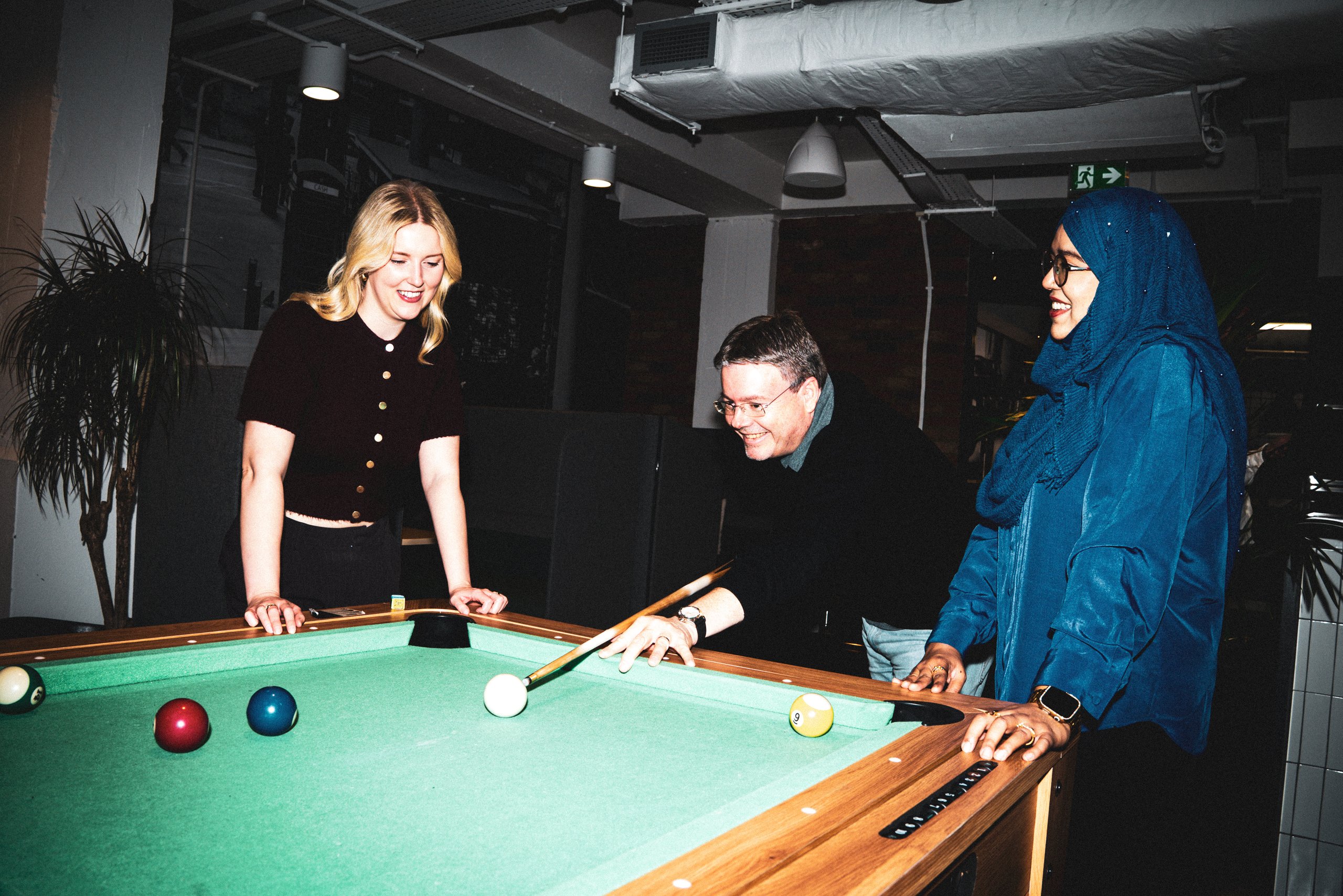 Three friends laughing while playing a game of pool in a casual, dimly lit venue.