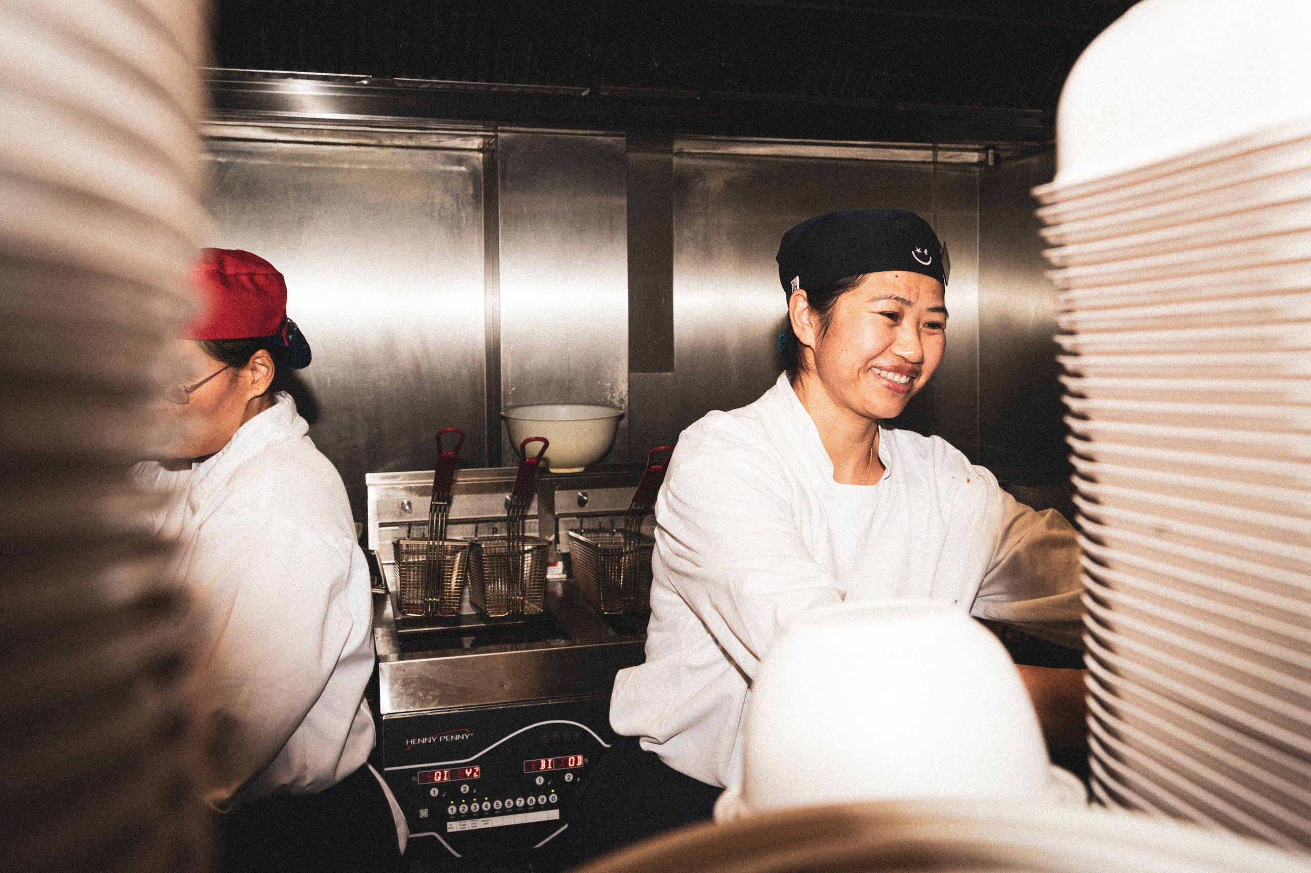 Two female kitchen workers wearing white uniforms and hats smiling while working in a fast food kitchen.