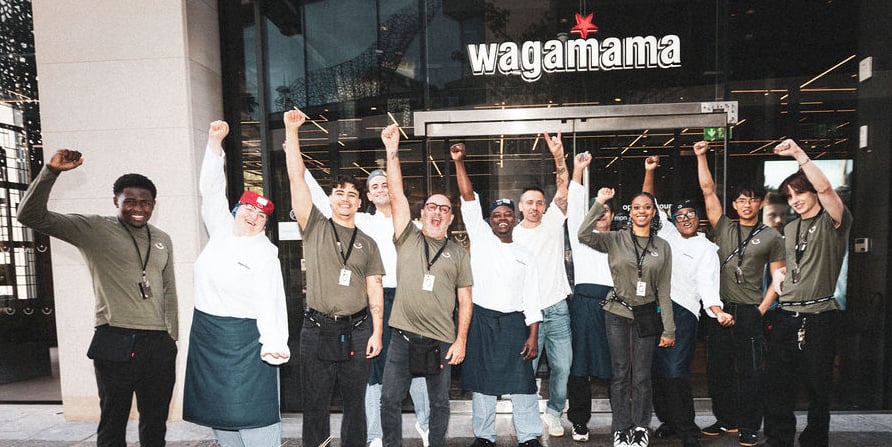 A group of Wagamama restaurant staff cheering with arms raised in front of the building entrance.