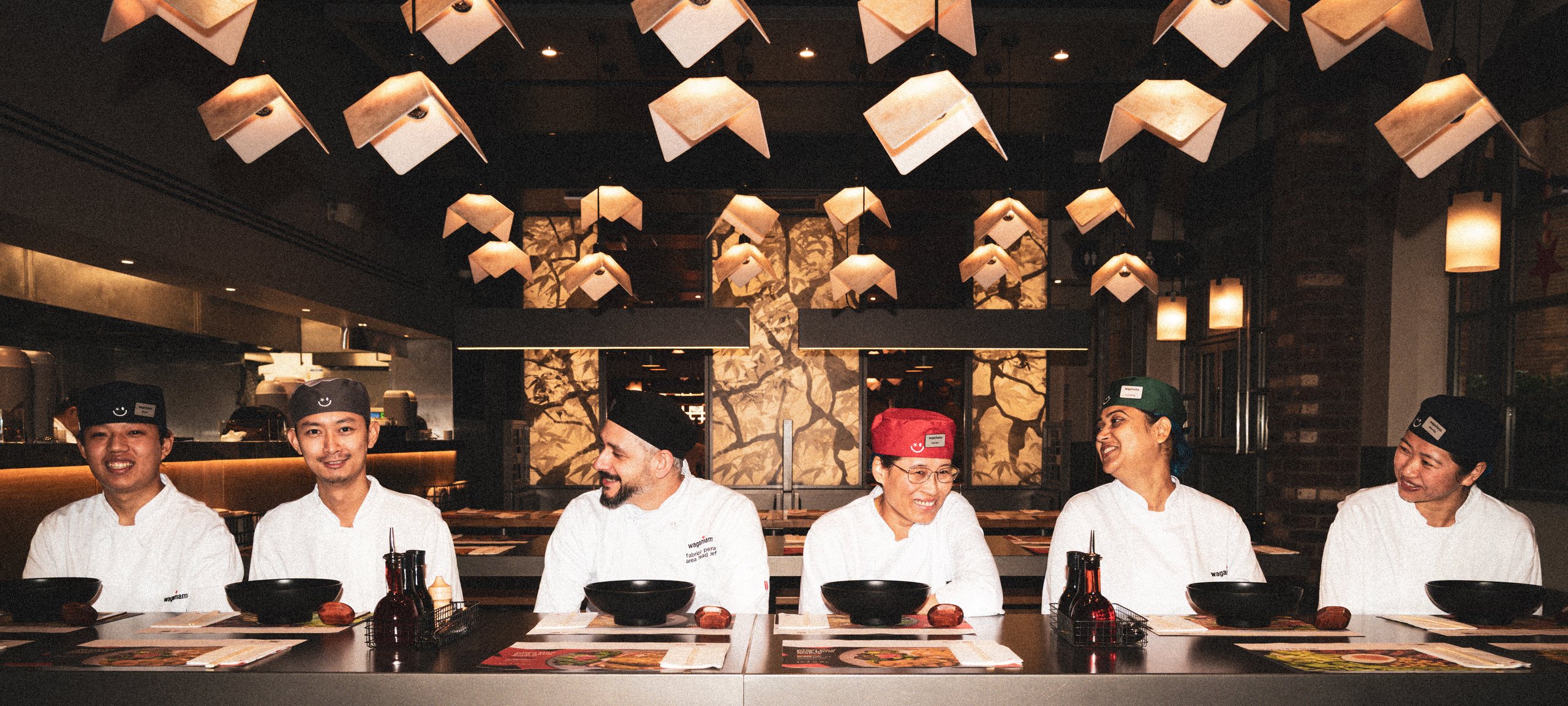 Six chefs in uniforms sitting at a long table in a restaurant with modern geometric lighting.