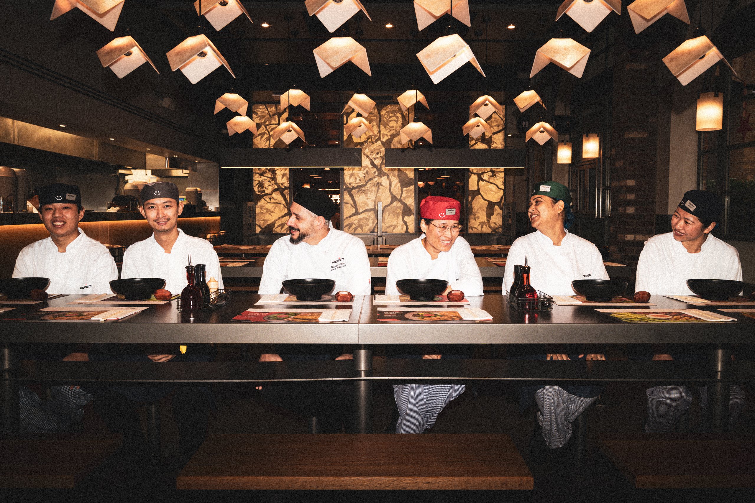 Six chefs in uniforms sitting at a long table in a restaurant with modern geometric lighting.