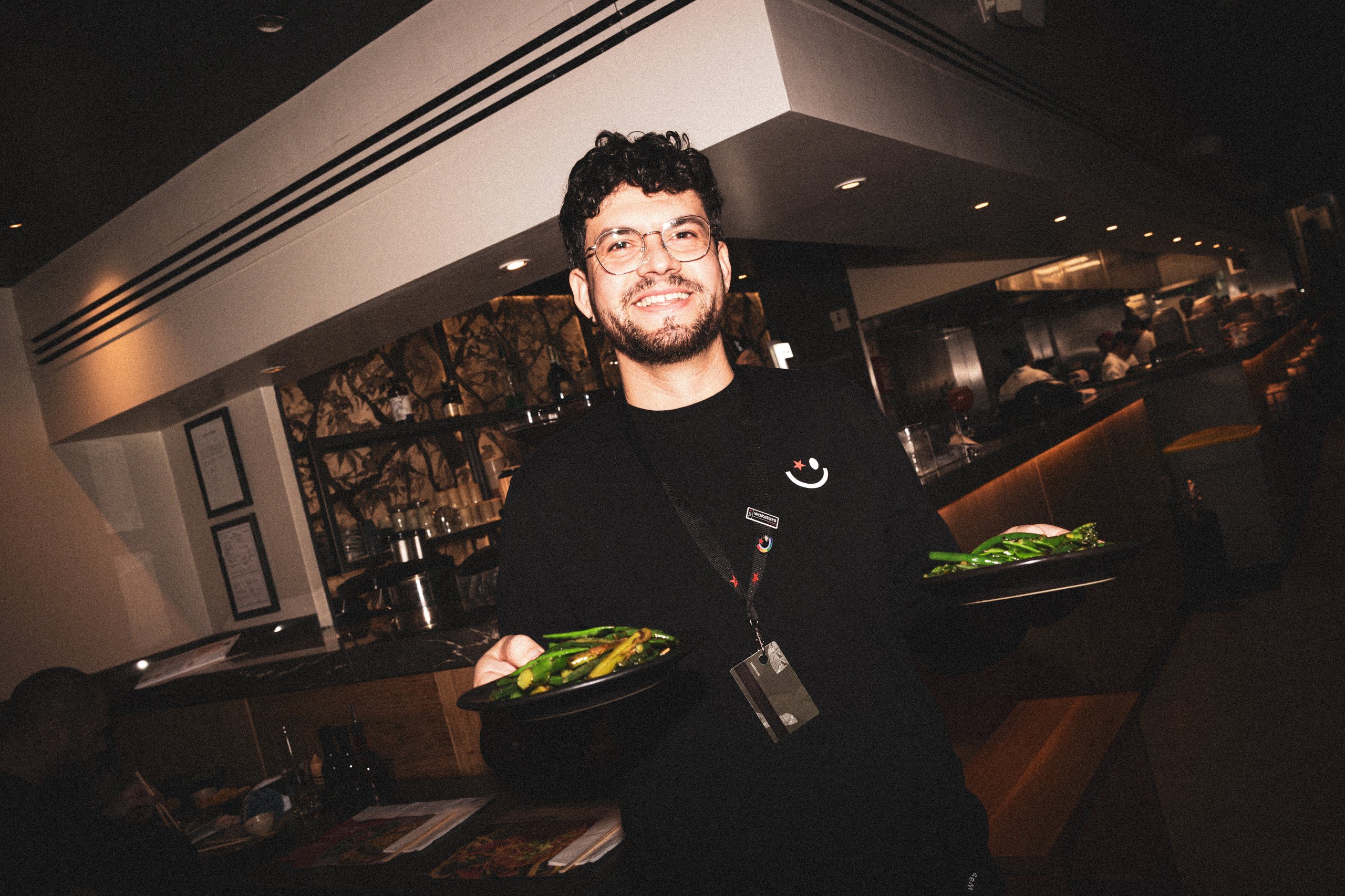 A smiling server in a black uniform carrying two plates of green beans in a restaurant.