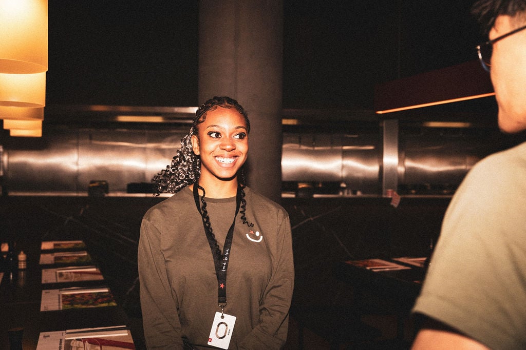 A smiling young woman in an olive green shirt and staff lanyard converses with a coworker.