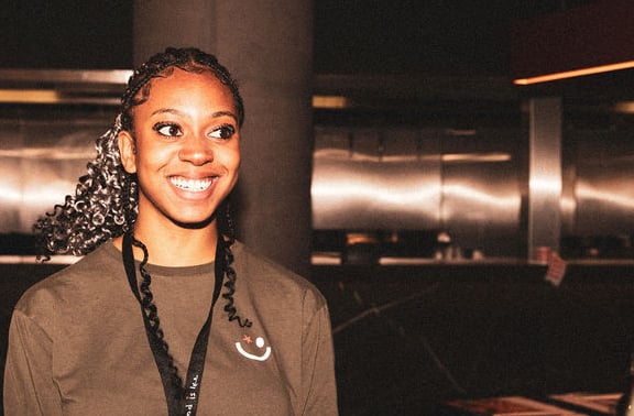 A smiling young woman in an olive green shirt and staff lanyard converses with a coworker.