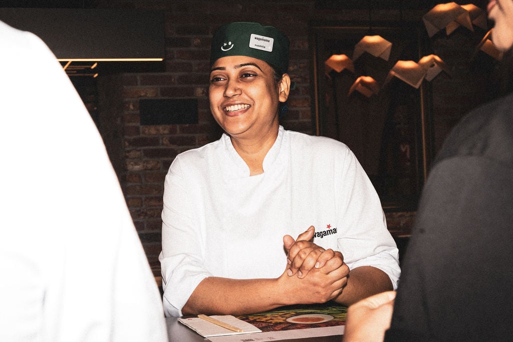 A smiling Wagamama restaurant chef wearing a green cap and white uniform sits at a table.
