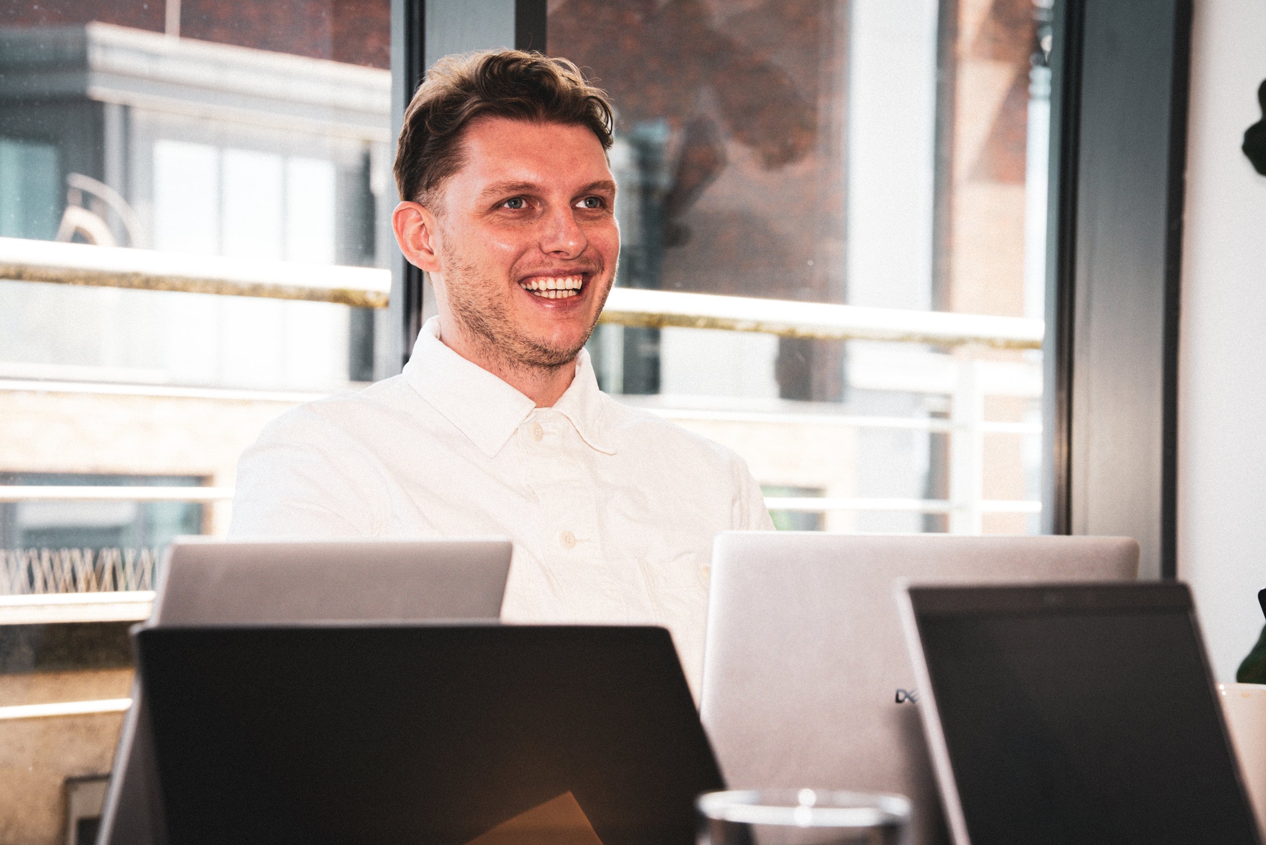 A smiling young man wearing a white shirt, sitting at a desk in an office with laptop computers.